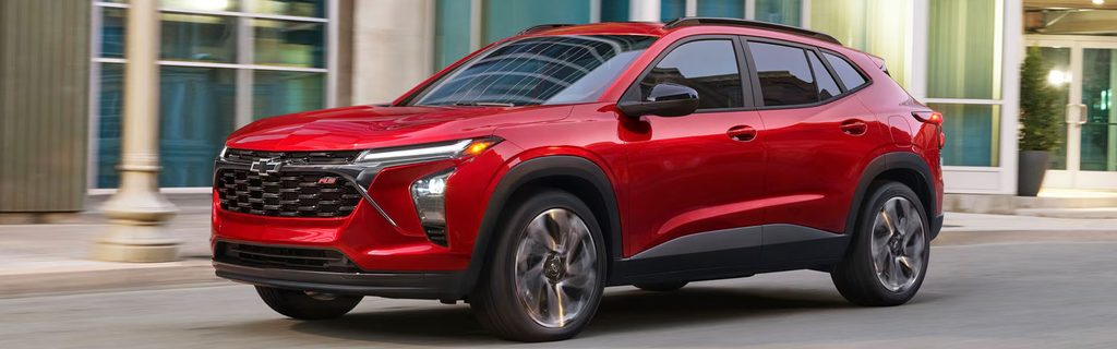 Red Chevrolet Trax SUV driving through a city street with modern buildings in the background.