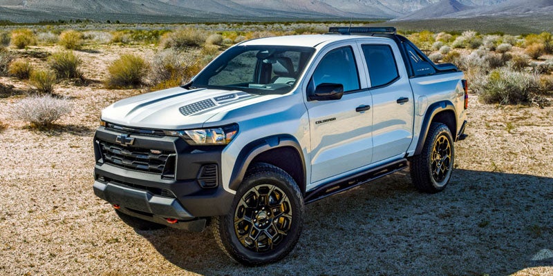 Silver Chevrolet Colorado parked on rocky desert terrain with mountains in the background.